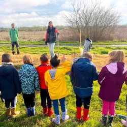 Enfants en bottes colorées écoutant deux animateurs dans un champ extérieur, le ciel est nuageux.