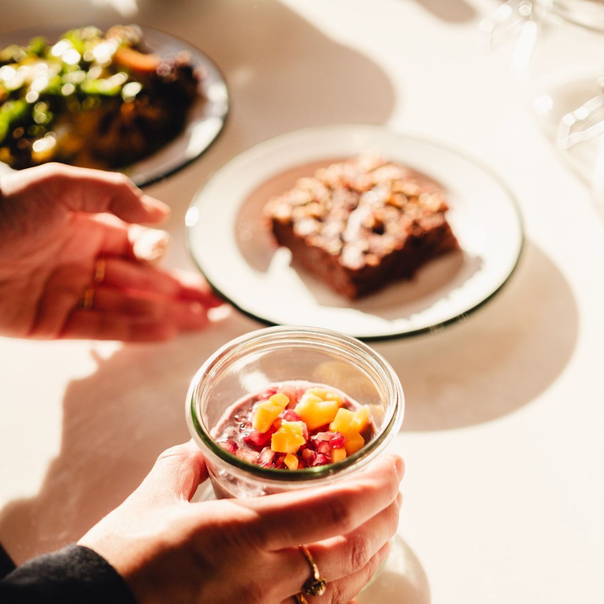 Person holding a glass jar with a layered dessert topped with diced fruit on a sunlit table with plates of food.