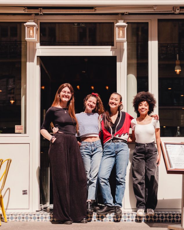 Four young women standing together outside MÛRE restaurant entrance, smiling and posing in casual attire under natural light.