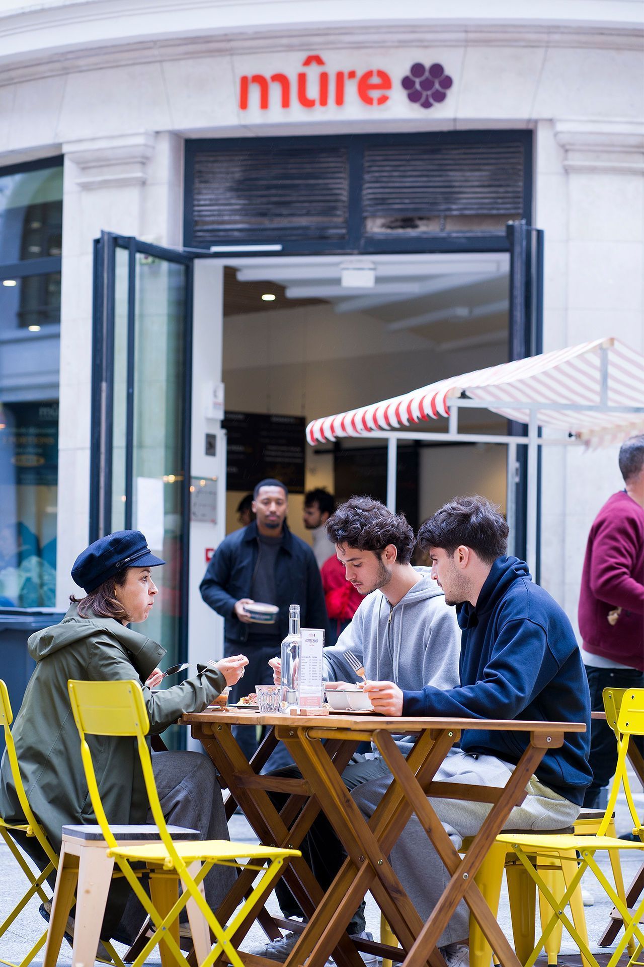 Trois clients dégustent un repas en terrasse sur des chaises jaunes devant le restaurant bio MÛRE à Paris.