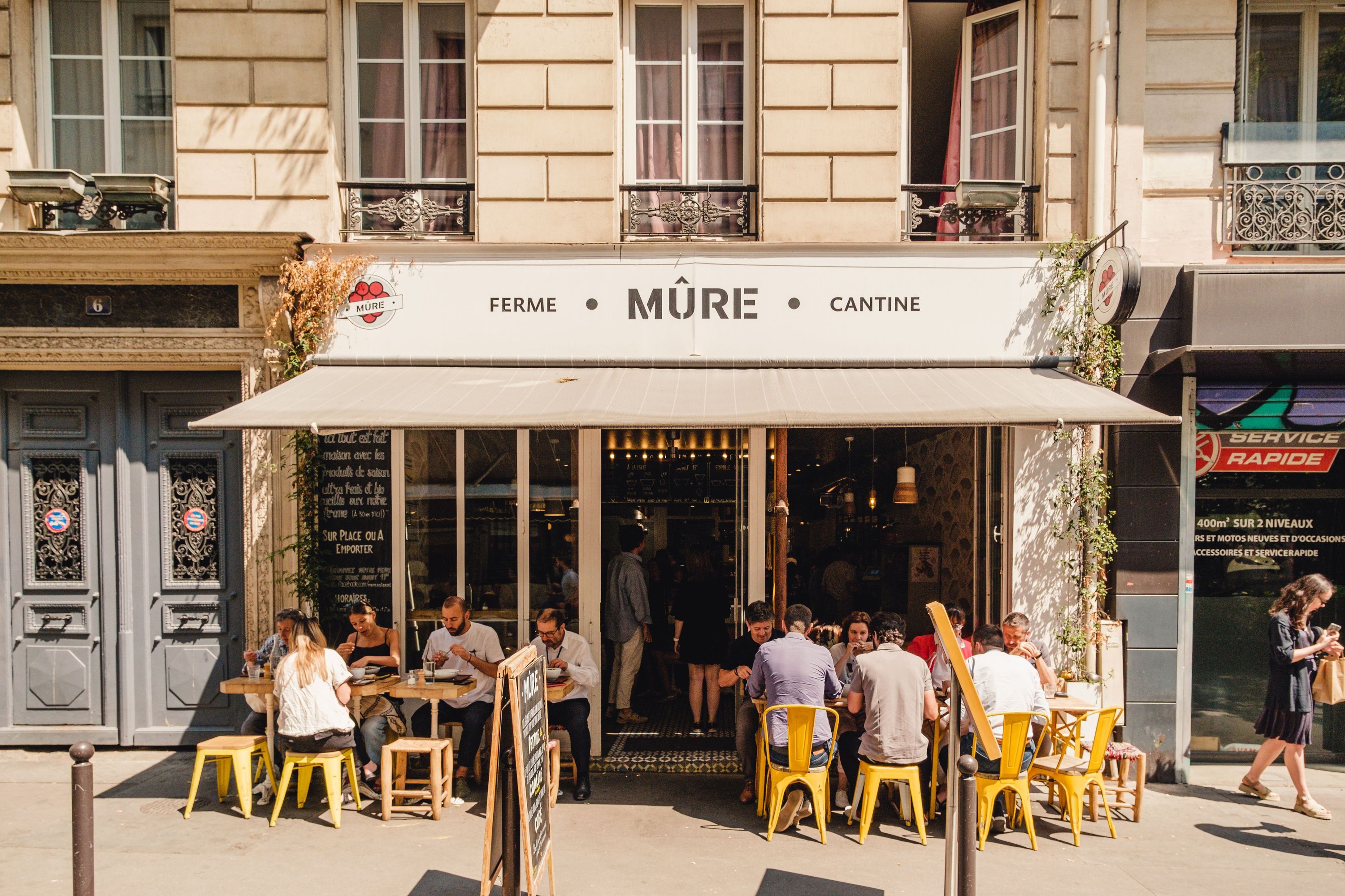 Terrasse extérieure du restaurant MÛRE avec des clients assis à des tables jaunes sous un store beige.