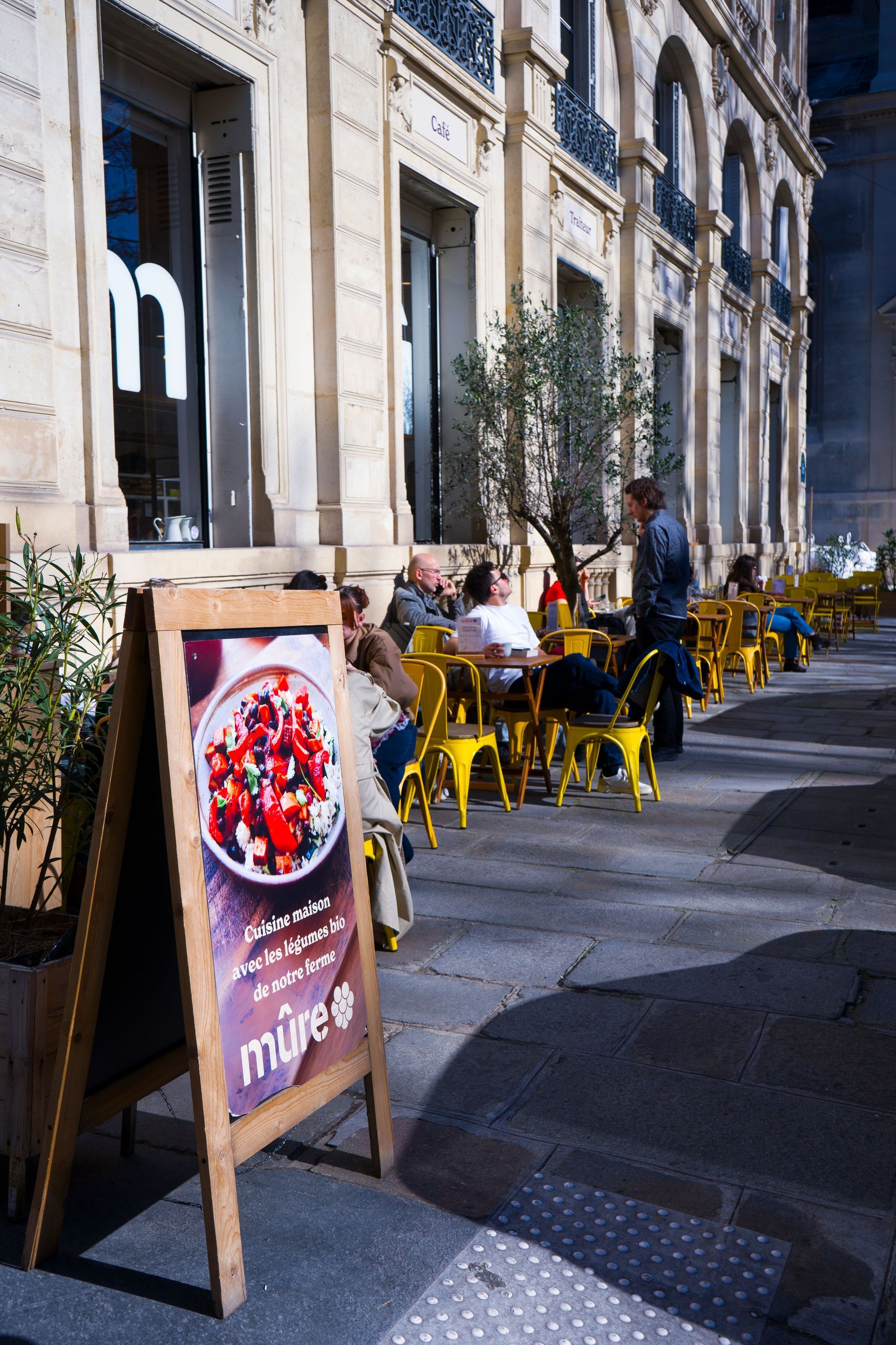 Terrasse de restaurant bio MÛRE avec tables, chaises jaunes, clients et panneau promotionnel à Paris.