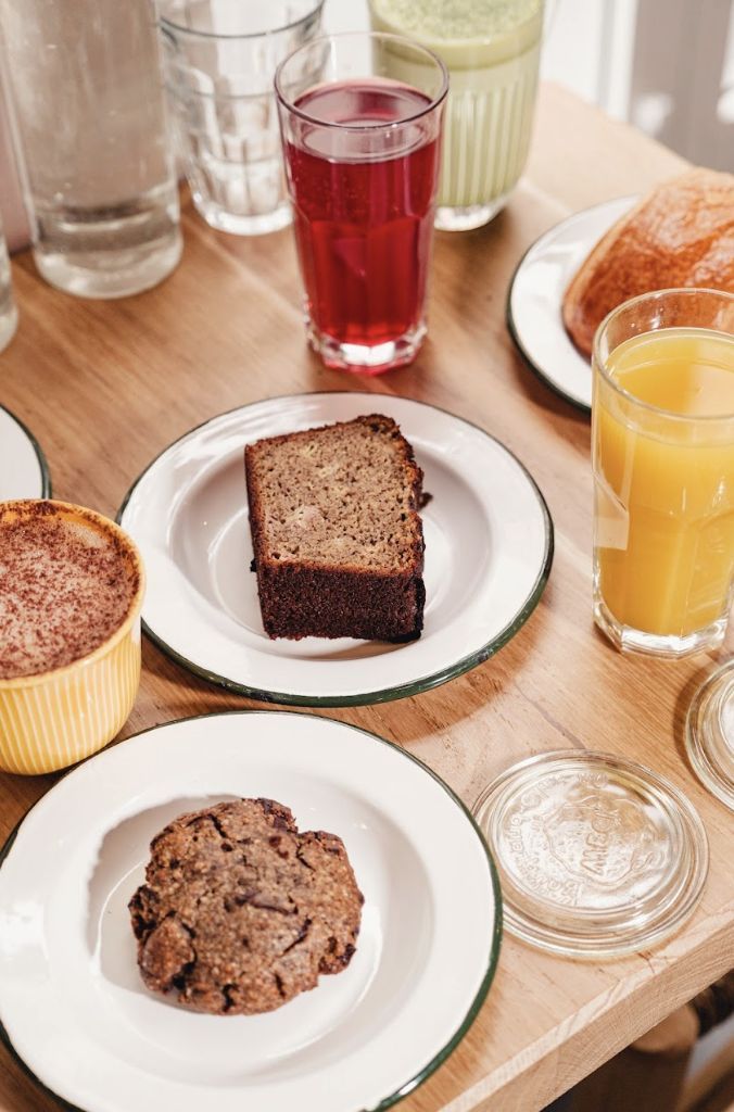 Various drinks and baked goods including cookies and cake slices are served on white plates on a wooden table at MÛRE.