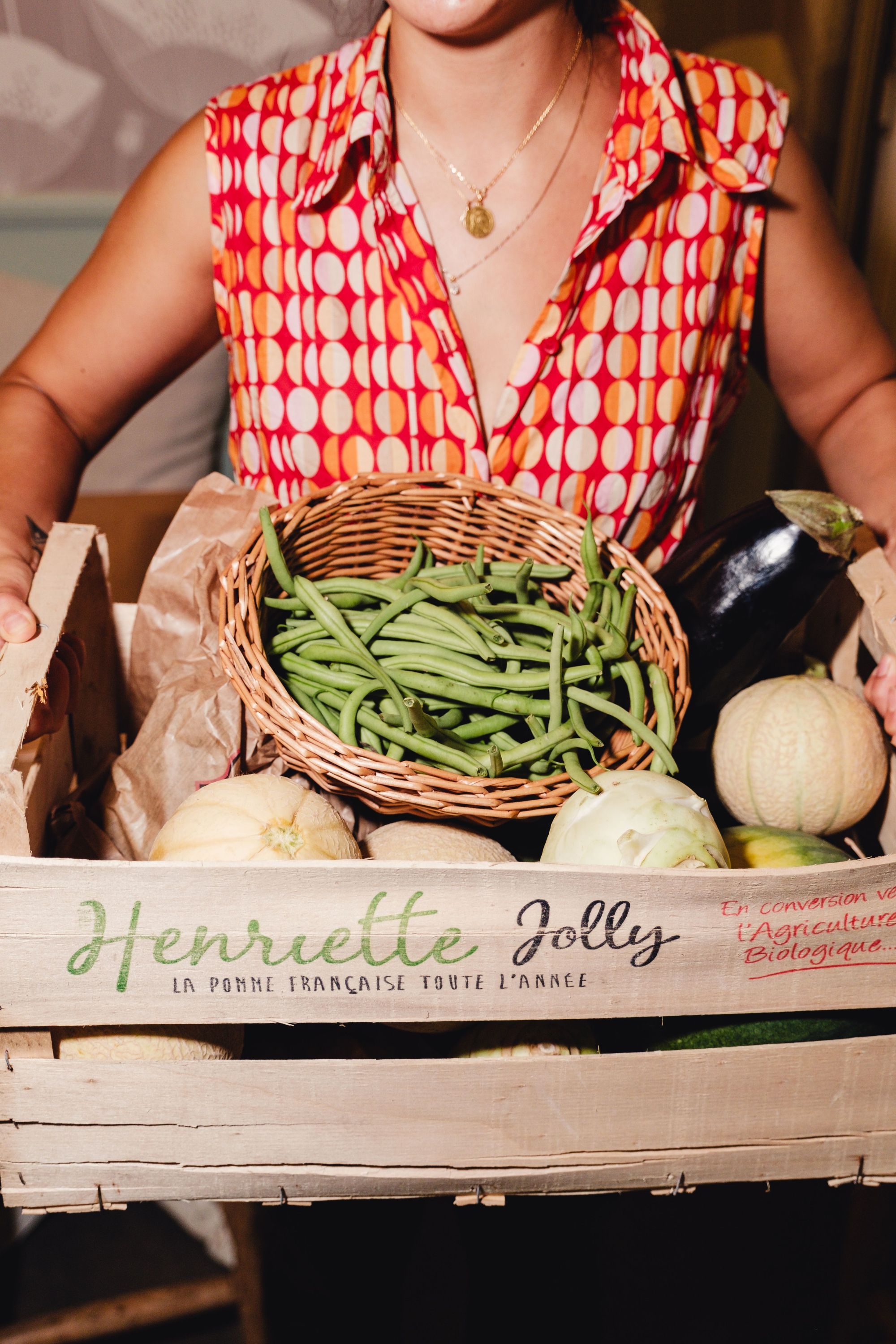 Panier en bois de légumes bio avec haricots verts, melons et aubergines, tenu par une personne en chemise à motifs colorés.