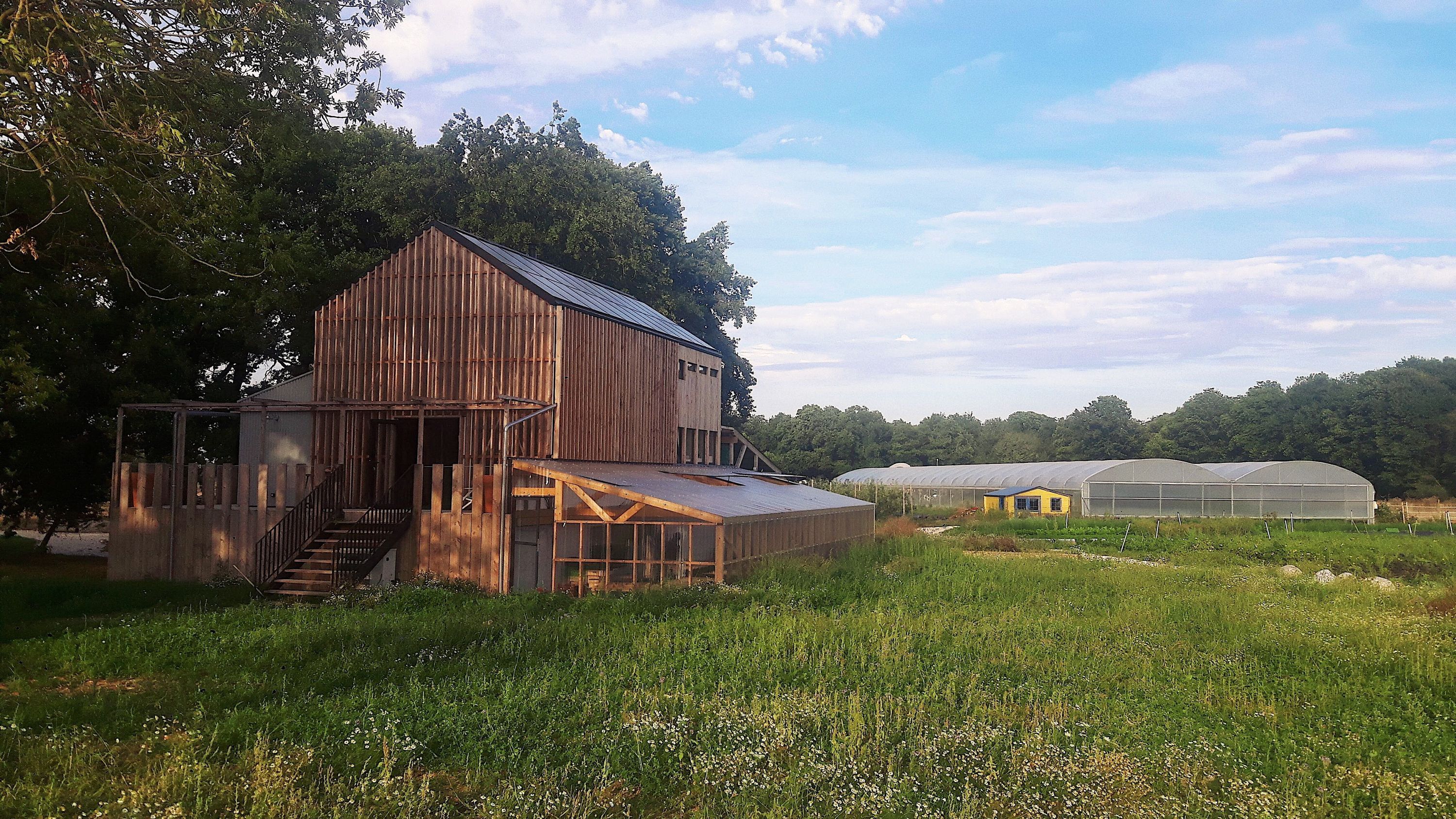 Ferme biologique avec serre et bâtiment en bois entourée de prairie verte sous un ciel partiellement nuageux.