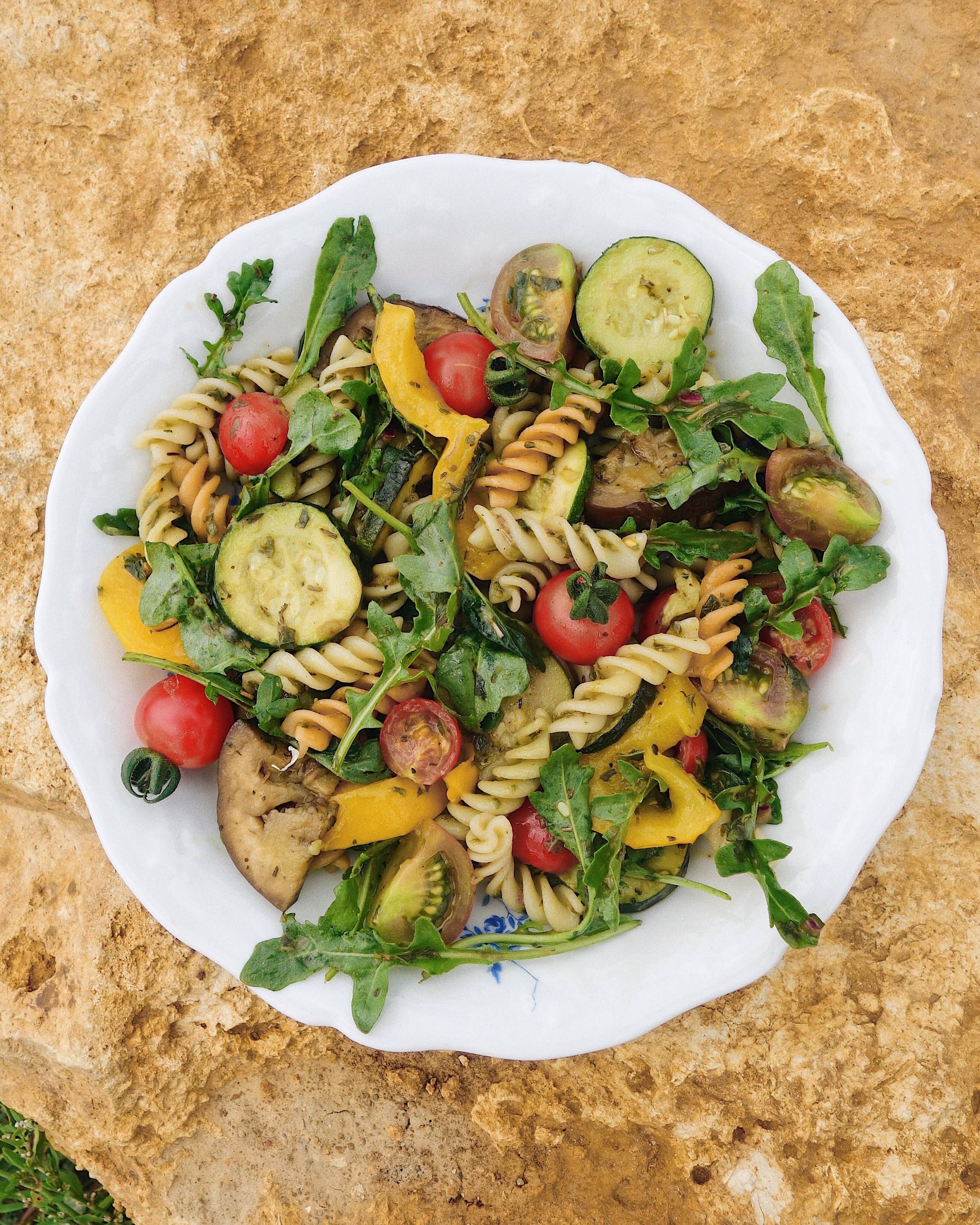 Fresh vegetable pasta salad with cherry tomatoes, zucchini, eggplant, yellow peppers, and arugula on a white plate.