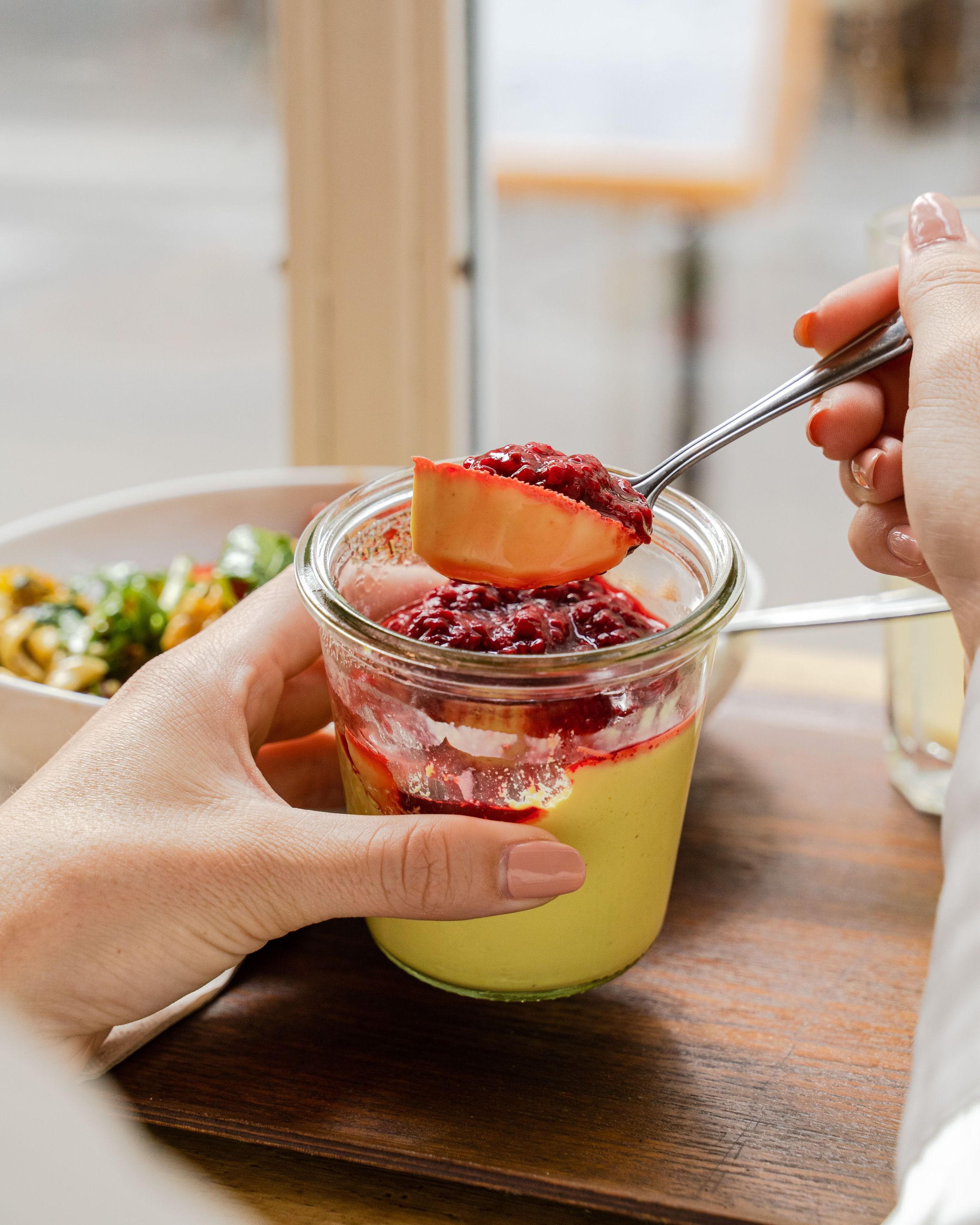 Dessert en verrine avec une couche de crème pistache et un coulis de fruits rouges tenue à la main sur une table en bois.
